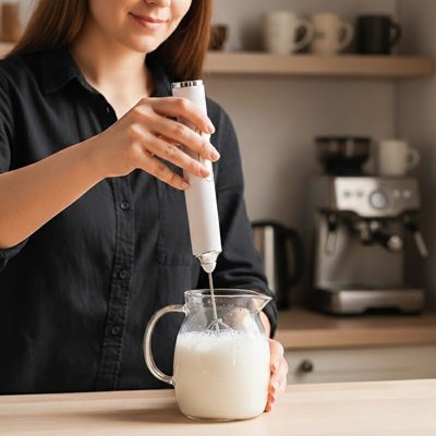 Mousseur à lait électrique en train de créer une mousse de lait dans un pichet tenu à la main dans une cuisine lumineuse.