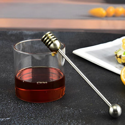 Cuillère à miel métallique posée sur un verre de miel avec fruits en arrière‑plan.