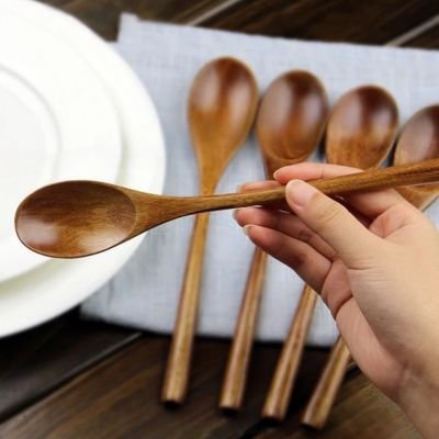 Cuillères en bois artisanales posées sur un linge gris, avec une cuillère tenue en main devant des assiettes en céramique.