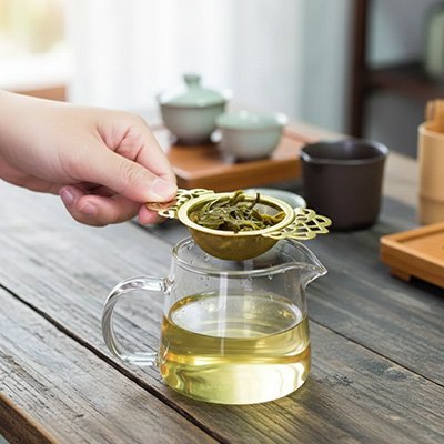 Passoire à thé en métal contenant des feuilles tenue au‑dessus d’une théière en verre.
